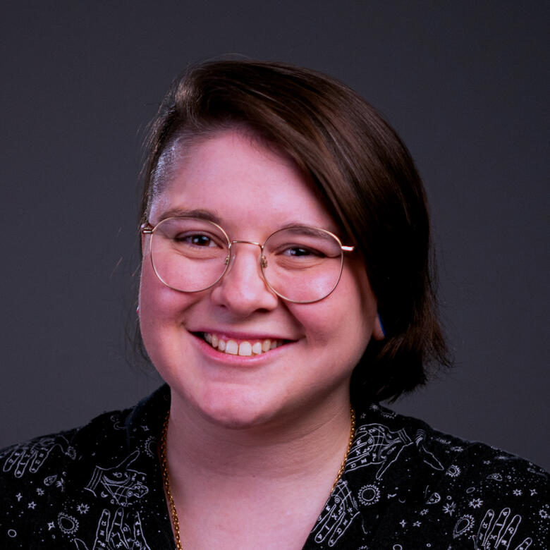 A photograph of a person with a glasses. They are wearing a black button down shirt, with one side of their head shaved, and the other side with hair to their chin. They are smiling at the camera, facing head on.